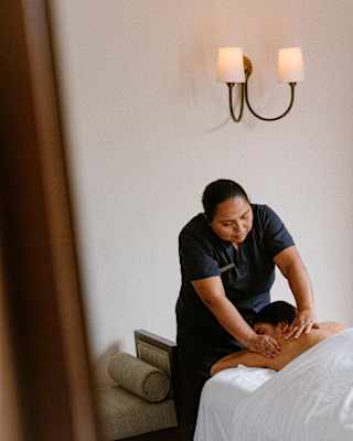 A female spa therapist in a dark uniform massages the back of a guest who lies face-down on a treatment bed beneath a sheet.