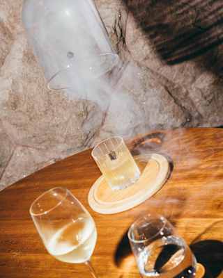 Tilted image of a waiter lifting a glass cloche to reveal a smoking pisco sour-hued cocktail, seen at a table with wine and water.