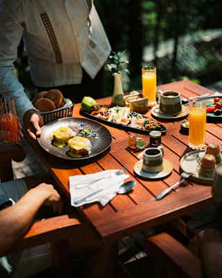 A waiter places a plate of eggs hollandaise on a sunny breakfast table with dishes of fruits, boiled eggs, coffee and juice.
