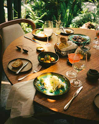 A beam of sun glances over plates of vibrant food at a table with wine and cocktails at Tampu Restaurant, seen from above.