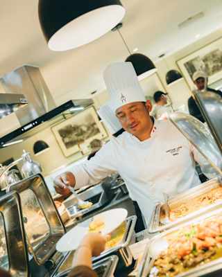 Angled image of a chef serving food from a buffet onto a plate held out in a hand, seen across a pass lit by a pendant light.