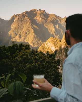 A man in a blue shirt stands with a drink on a viewing shady platform, looking out at the sun-capped contours of the Andes.