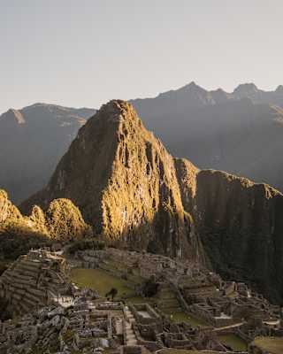 Green-gold with sun, Huayna Picchu looms over the shaded Machu Picchu ruins in a breathtaking aerial view of the citadel.