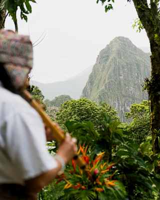 A local shaman wearing a woven hat plays flute facing a view of Huayna Picchu, rising against a cloudy sky.