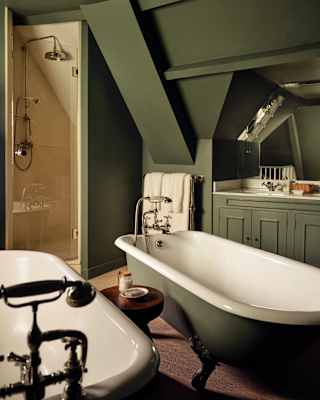 A shower fits under the eaves of the Botticelli Room bathroom, seen over twin tubs, painted dark olive, matching the décor.