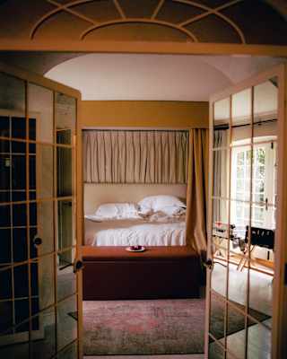 View though double doors, with mirrored panels, to a bedroom, where light from a window falls onto a white unmade bed.