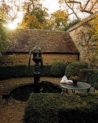 Ancient stone walls and trimmed box hedges surround a small fountain. A book lies open on the ironwork garden table