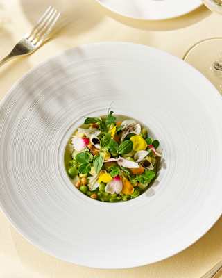 A wide-brimmed white bowl cradles a risotto of garden vegetables and herbs with vibrant bursts of green, yellow and pink.