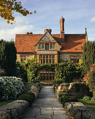 View of Le Manoir Aux Quatre Saisons, stone aglow in honey sunlight, seen from a paved garden path bordered by lush shrubs.