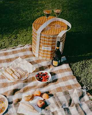 Two flutes sit on a cylindrical orange wicker hamper on a picnic blanket with cherries, sandwiches and a Champagne bottle.