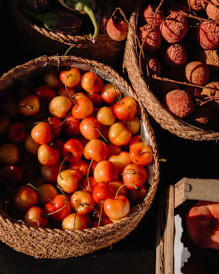 Looking down on wicker baskets of rosy crab apples and lychees, with a glimpse of a peach in a nearby crate, seen from above.