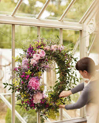 A woman creates a joyful summer wreath with pale pink Peonies, blushing Roses and deep cerise Asters on a ring of leaves