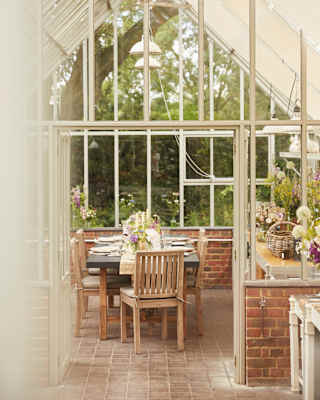 View through open doors into the glasshouse, where a laid table with floral displays awaits guests in the light-filled space.