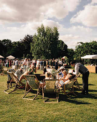 Guests sit on floral deckchairs and mill around the spacious lawn at a summer garden party with gazebos of refreshments