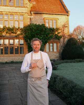 Chef Arnaud Donckele stands on a path in front of Le Manoir, smiling to camera and holding spectacles, face in late sun.