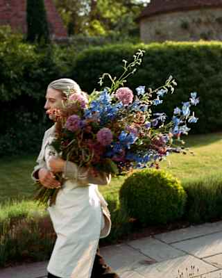A member of staff in a white apron carries an enormous bouquet of pink alliums and blue amsonias, picked from the garden.