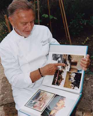 Chef Raymond Blanc, wearing chef whites, smiles as he points to old photographs in a photo album.