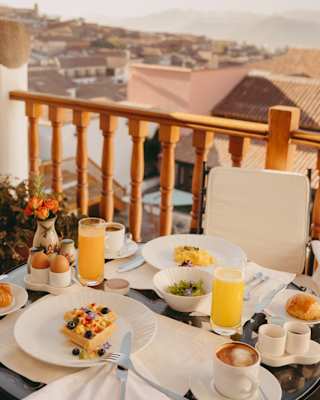 Early sun glows on a spread of waffles, eggs, orange juice and coffee on a balcony table by wooden railings, with city views.