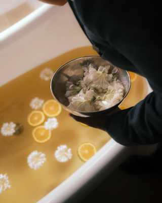 A woman leans over a bath with a bowl of white blooms to add to the flowers and lemon slices afloat in the honey-hued water.