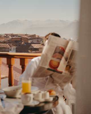 In soft-focus foreground, a guest reads at a breakfast table on a balcony with city views, face hidden by an open newspaper.