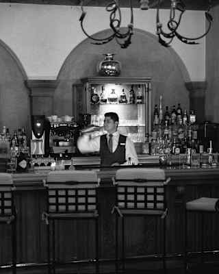 A barman shakes a cocktail behind the bar at Senzo Bar, where four vacant high-backed stools await guests, in monochrome.