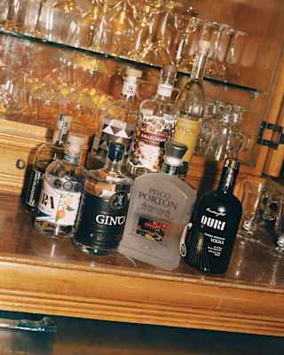 Tilted image of bottles of spirits grouped together on a wooden counter in front of shelves of wine and cocktail glasses.