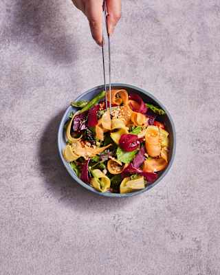 A hand adds ingredients with tweezers to seasonal salad with strips of bright root vegetables and leaves, seen from above.