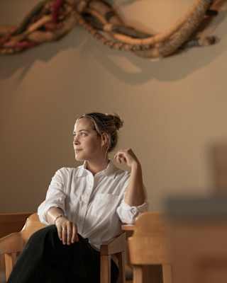 Soft light illuminates Mauka Chef Pía León who sits in her restaurant in a white top with black trousers and hair tied back.