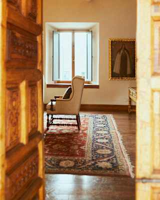 A white armchair sits close to a light-filled window on a vintage woven red rug in a suite, viewed through an open wood door.