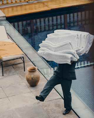 A staff member tips backwards, carrying a large pile of folded, striped towels at the edge of the pool, seen from above.