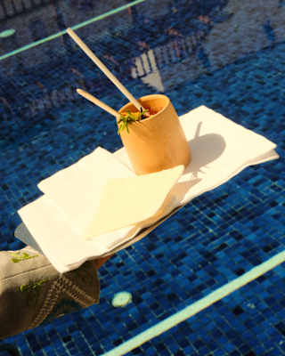 A waiter's arm carries a yellow tumbler with a drink, two stirring sticks and a rosemary garnish, on a tray by the pool.