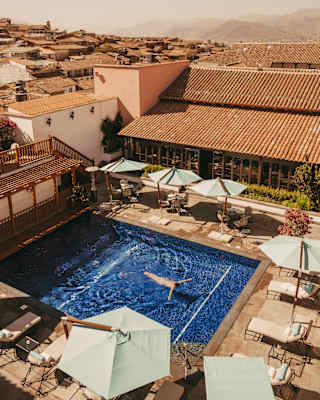 A guest floats in the azure pool, lined with parasols in a courtyard of terracotta-tiled buildings, seen from above.