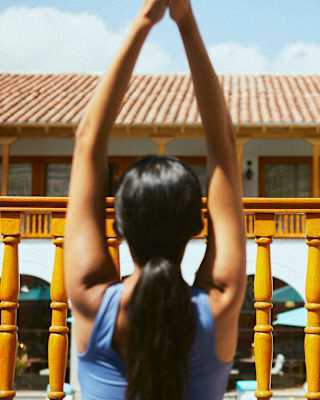 In soft-focus, a woman in a blue top with long dark hair holds up her arms as she does yoga on a loggia, seen from behind.