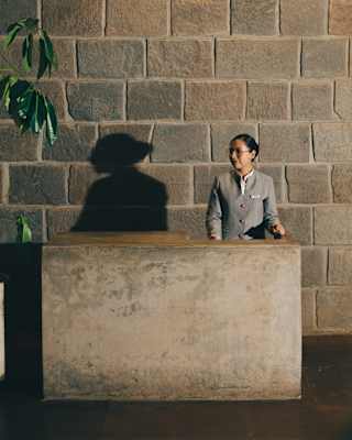 A female receptionist with neat dark hair, glasses and grey suit casts a profile shadow on the wall behind a stone desk.