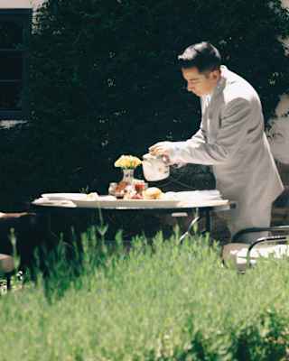A male member of staff in a grey suit pours tea from a white teapot at a table laid for breakfast in the sun, seen over a hedge.