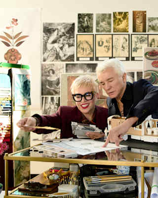 Baz Luhrmann, in black, and Catherine Martin in burgundy with glasses, look through designs in a busy art studio.