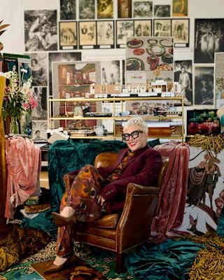 Catherine Martin, in a burgundy jacket over a deep orange-print playsuit, sits in a leather chair among fabrics in a studio.
