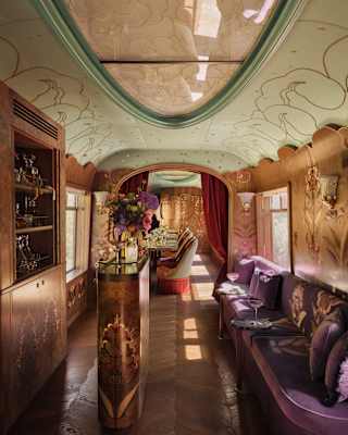 Luxurious vintage train carriage interior with ornate wood panelling, velvet seating, floral arrangements, decorative ceiling panels, and soft ambient lighting. A long table is visible further down the carriage.