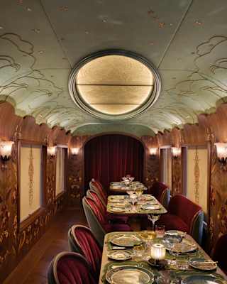 An elegant, vintage-style train dining car with plush red velvet seats, ornate wooden panelling, set tables with fine china and glassware, and a large oval ceiling light.