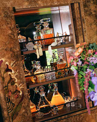 A glass cupboard filled with various crystal decanters and glasses, some containing amber liquid, is displayed. Decorative flowers and hanging ornaments are also visible near the cupboard.