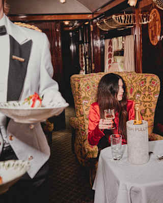 A waiter walks by woman with long dark hair and red satin top as she sits at an Ibis carriage table drinking Veuve Clicquot.