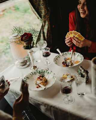 Two guests play cards as they enjoy bowls of desserts at a table with red wine and water, with a bud vase decoration.