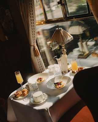A small table set for breakfast by a sunlit window, featuring plates of food, croissants, drinks, glasses, and a lamp, all on a white tablecloth with elegant, cosy decor.