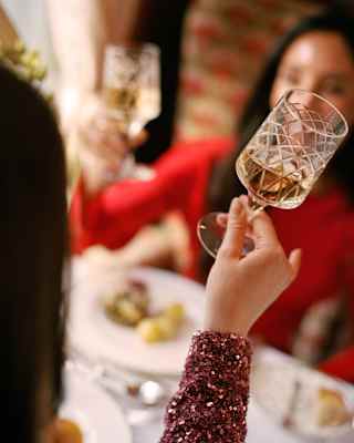 A woman with a purple sequined sleeve, mainly off-camera, raises a drink for a toast, mirrored by a friend in red, opposite.