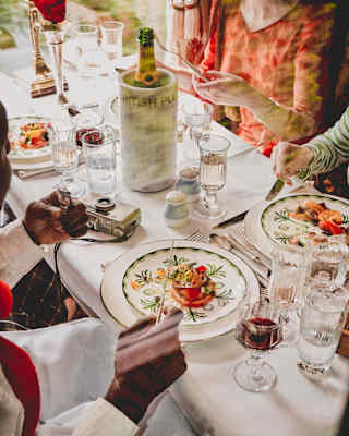 Four guests enjoy a vibrant lunch at a table busy with glasses of red wine, water and Champagne, seen from above, close-up.
