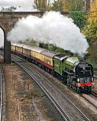 A plume of steam billows out along the length of the cream and burgundy carriages as the train passes below an arched bridge