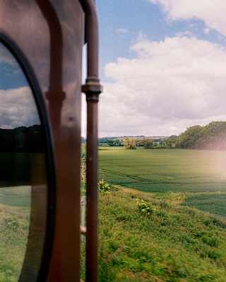 View of beautiful green crop fields bordered by forest beneath billowing white clouds, seen from an open train window.