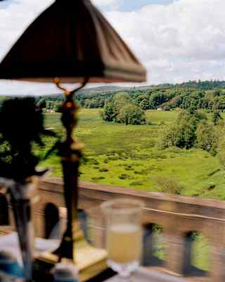 View of lush, undulating green fields bordered by forest, seen through a carriage window from a dining table with Champagne.