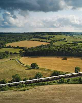 Gently rolling hills reveal a patchwork of yellow and green fields and woods. In the foreground the elegant steam train snakes