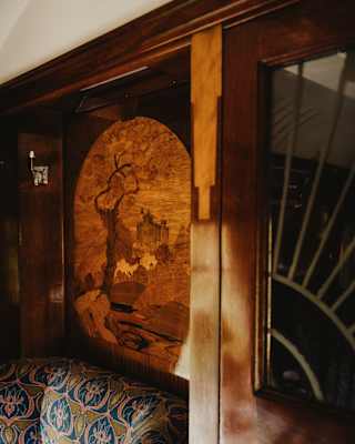 A wooden wall panel featuring an engraved tree and landscape scene is next to a sofa with patterned upholstery and a window with decorative glass. Warm sunlight highlights the cosy, vintage interior.